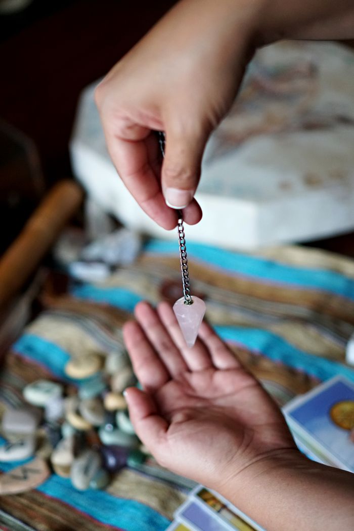 about-us Two hands using a rose quartz pendulum over tarot cards for divination purposes.