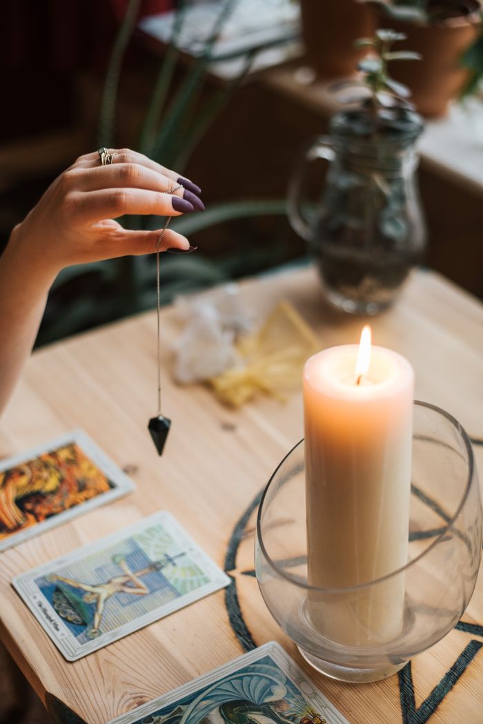 services-04 From above of crop anonymous female fortune teller with amulet on thread above tarot cards near burning candle on table