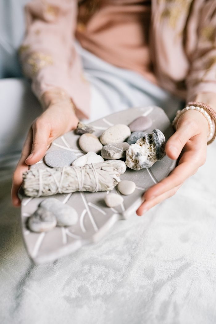 A calming scene with hands holding a sage bundle and stones on a decorative plate indoors.